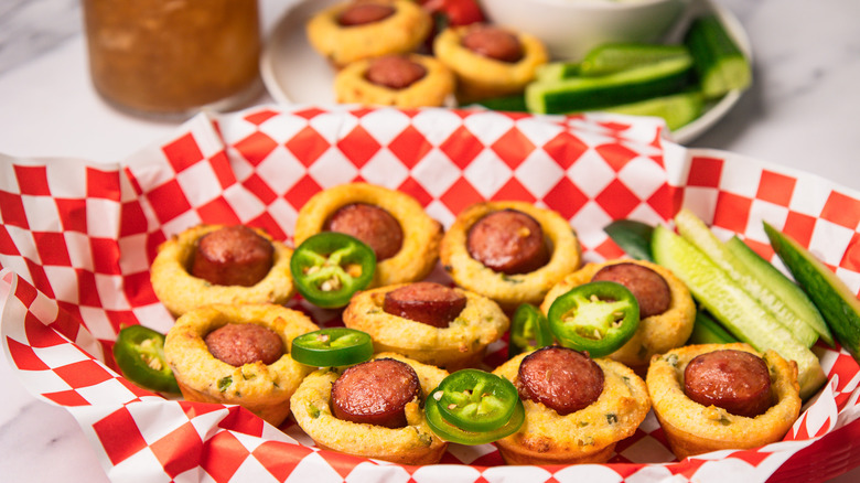 Mini corn dogs in basket with fresh cucumbers and jalapenos, and more on a plate in background.