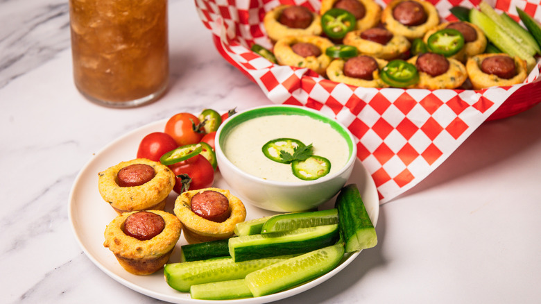 Mini corn dogs on plate with fresh cucumbers and jalapeno crema, with iced tea and more corn dogs in basket in background.