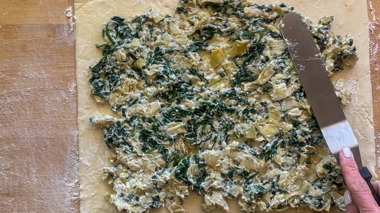 Spatula spreading spinach artichoke dip across puff pastry