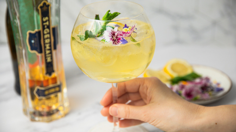Hand holding springtime Hugo Spritz with mint, edible flowers and lemon slice