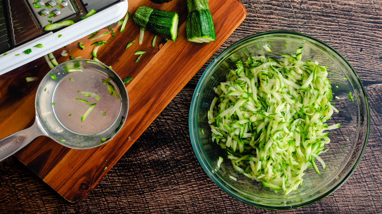 shredded zucchini in a glass bowl on a table near a cutting board