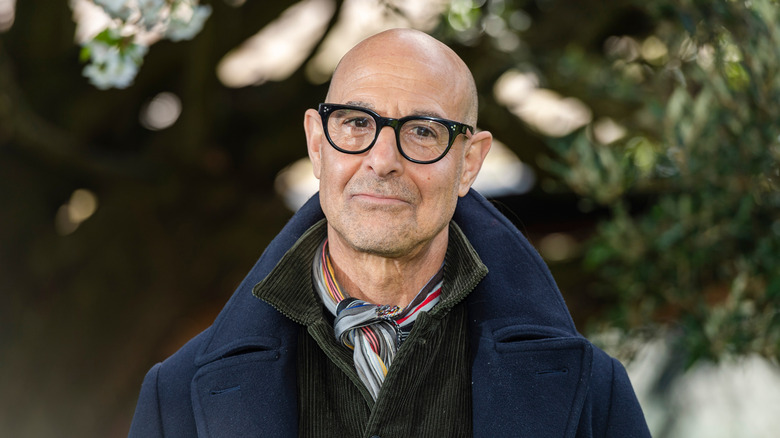 Smiling Stanley Tucci with black glasses, colored scarf, and blue coat