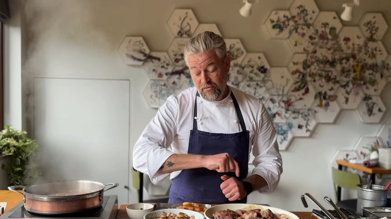 Chef Adam Byatt stands in a kitchen with several dishes containing ingredients for coq au vin, next to a simmering pot