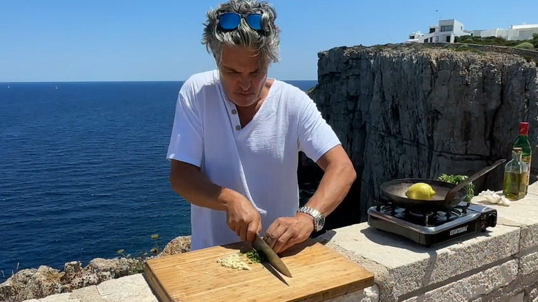 Peter Hanley of Pete's Pans dices vegetables next to a pan on a portable stove, with the ocean and a cliff-face in the background