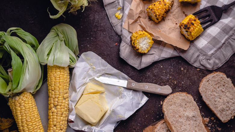 Uncooked corn on the cobs, an opened packet of butter, some bread slices, and some grilled corn on the cob pieces