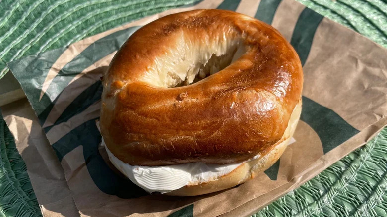 A plain Starbucks bagel with cream cheese sitting on a paper bag in the sun