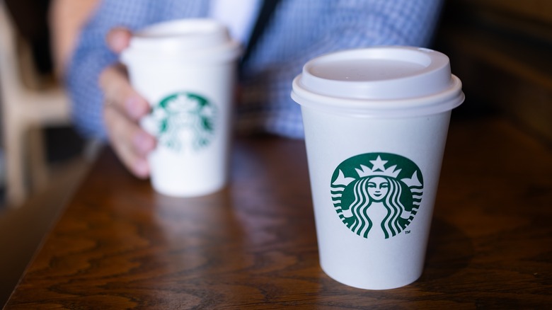 Two Starbucks drinks on a wooden table, one being held by a person in a blue shirt