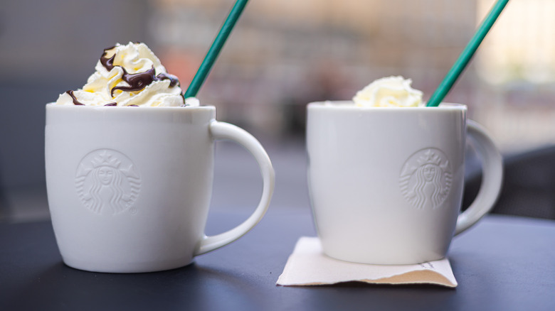 Two white mugs with Starbucks logo holding drinks with whipped cream and green straws