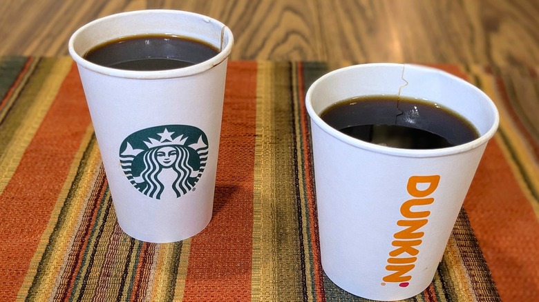 A paper Starbucks cup and a paper Dunkin' cup with black coffee on striped linen