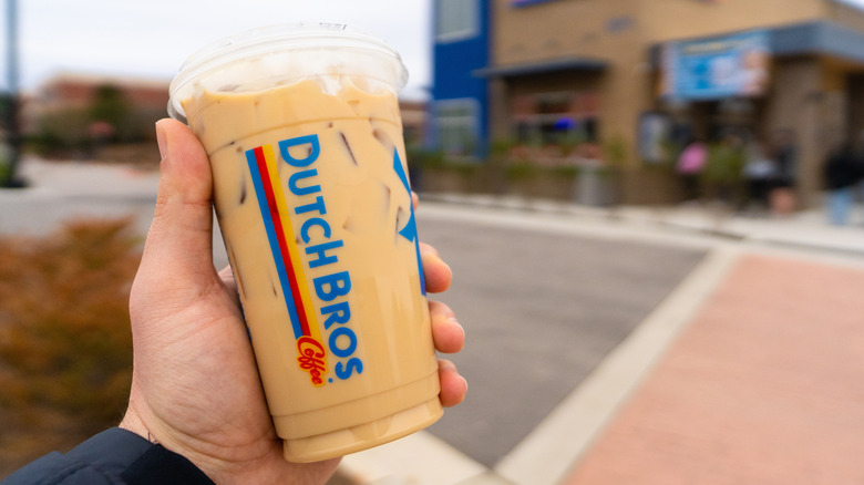 A hand holding a Dutch Bros drink in a plastic cup in front of a Dutch Bros location