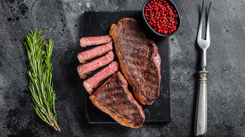 grilled, sliced steak with fat cap on a slate with pink peppercorns, a carving fork, and a sprig of rosemary