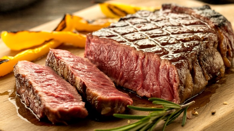 Partially sliced medium rare grilled ribeye steak on wooden board next to rosemary sprig and yellow peppers