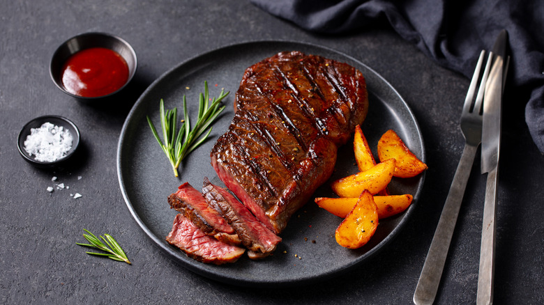 A medium rare grilled steak on a black plate with some golden potatoes and salt and ketchup to the side
