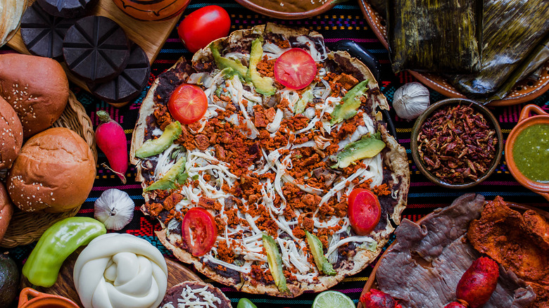 Tlayuda topped with beans, cheese, chorizo, avocado, and tomatoes surrounded by Oaxacan ingredients