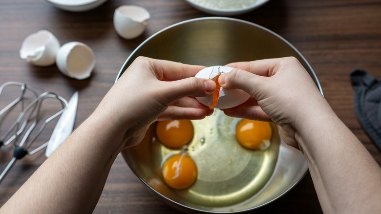 A person uses two hands to open a cracked egg over a bowl full of other raw eggs