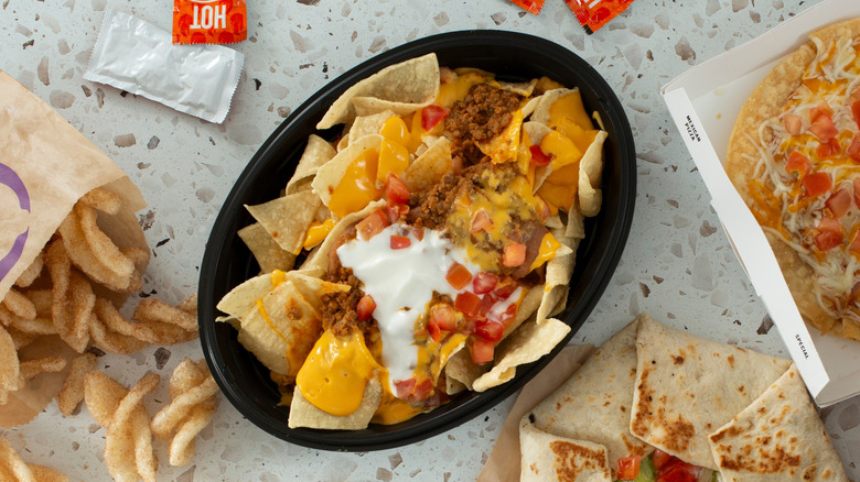an overhead view of a plastic bowl of Taco Bell nachos on a stone countertop beside hot sauce packets and other Taco Bell menu items
