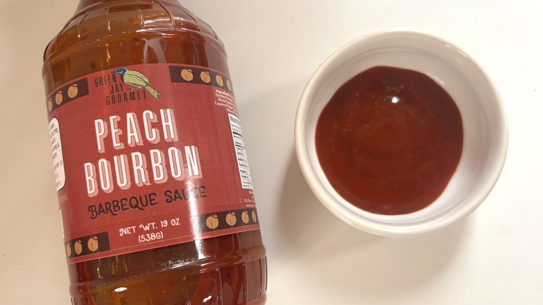 Bottle of Green Jay Gourmet Peach Bourbon BBQ Sauce next to small white dish of bbq sauce on white table