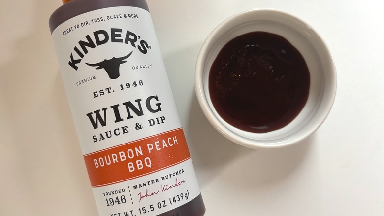 Bottle of Kinder's Bourbon Peach BBQ Sauce next to small white bowl of sauce on white table