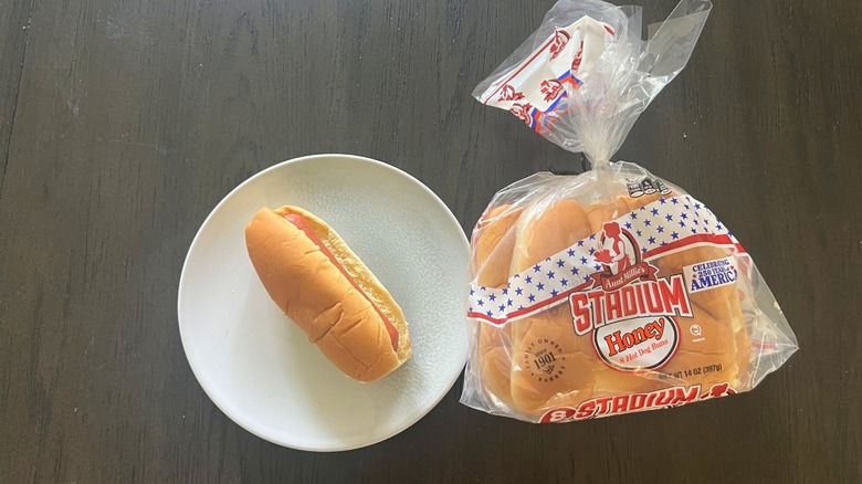 Aunt Millie's Stadium Honey Hot Dog Bun beg next to a hot dog served on a white plate