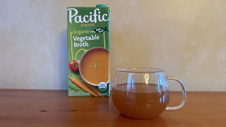 Carton of Pacific Foods vegetable broth on wooden table with glass of broth