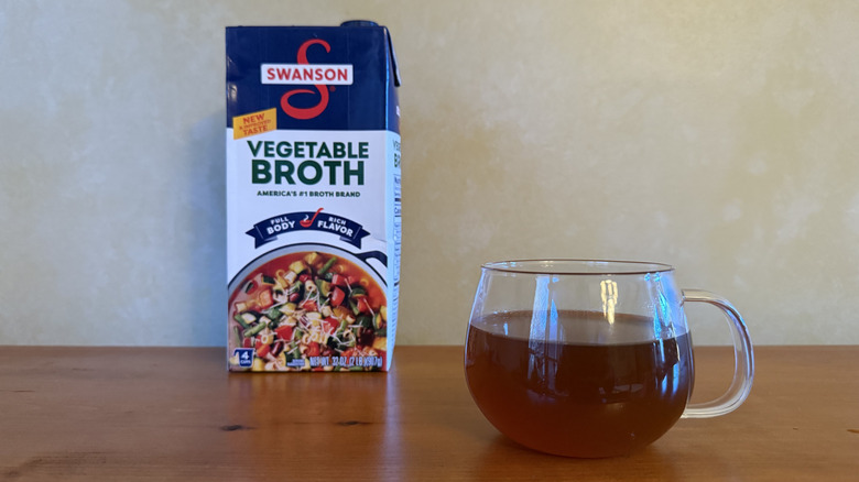 Carton of Swanson vegetable broth on wooden table with cup of broth