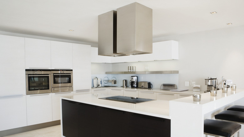 A sleek, white, modern kitchen with a large marble kitchen island that has two stainless steel range hoods mounted over it