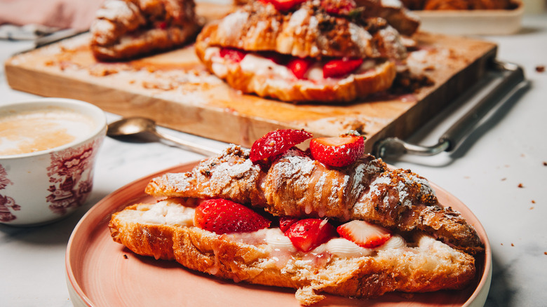 A strawberry cheesecake croissant on a plate besides more on a cutting board in background.