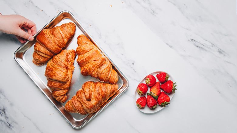 Four croissants on metal sheet besides strawberries.