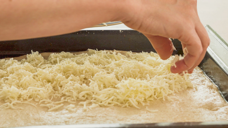 person sprinkling grated cheese on a pizza base on a sheet pan