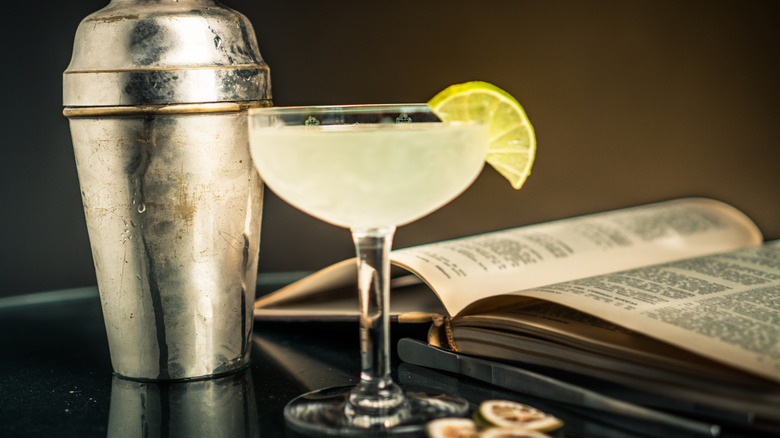 Cocktail shaker next to gimlet with lime wedge and book.