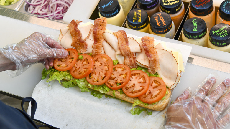 a gloved Subway employee assembles a sandwich with turkey, bacon, lettuce, and tomato