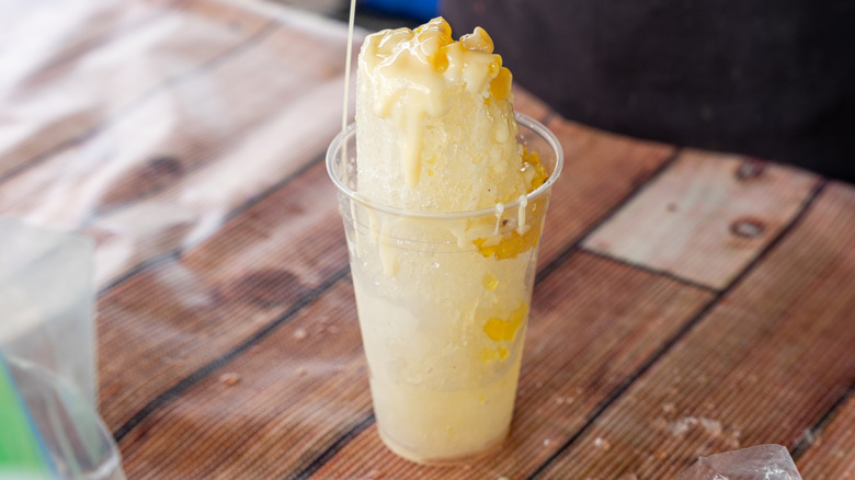A raspado topped with pineapple and condensed milk in a clear plastic cup on a faux wood table