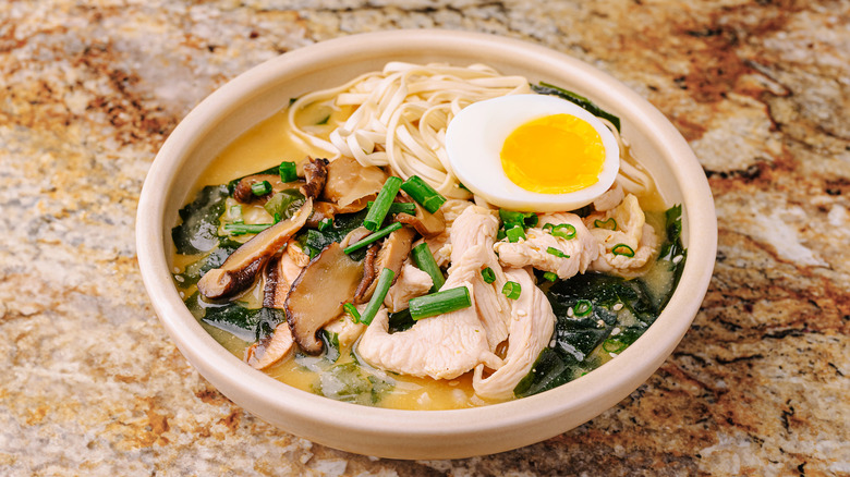 A bowl of ramen sitting on a table top. The bowl is filled with mushrooms, noodles, chicken, scallions, and half a soft-boiled egg