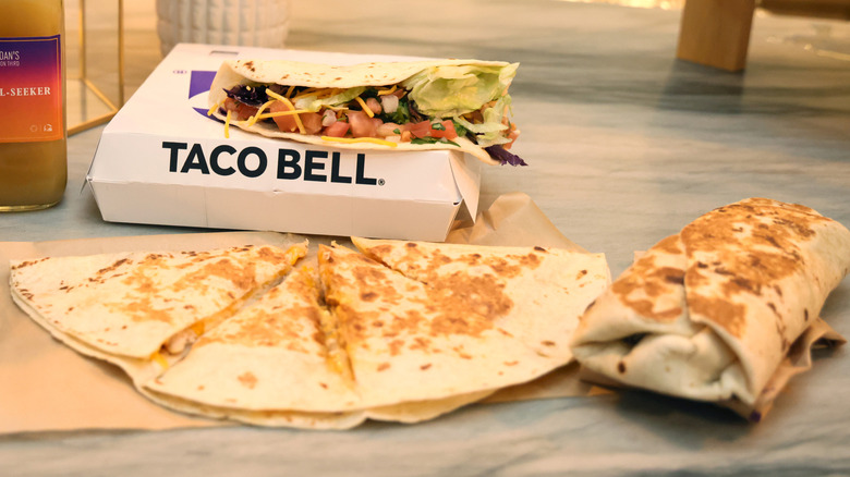 A quesadilla and a grilled burrito on a table with a Taco Bell box behind them and a soft taco sitting a top it