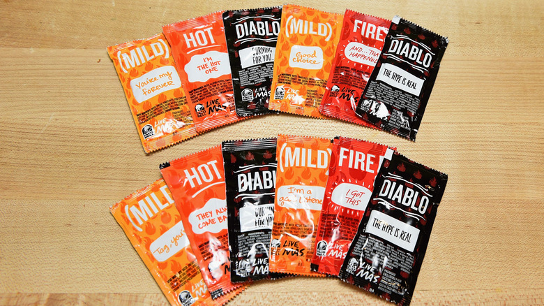 Two rows of Taco Bell sauce packets fanned out on a wooden table featuring mild, hot, fire, and Diablo sauces