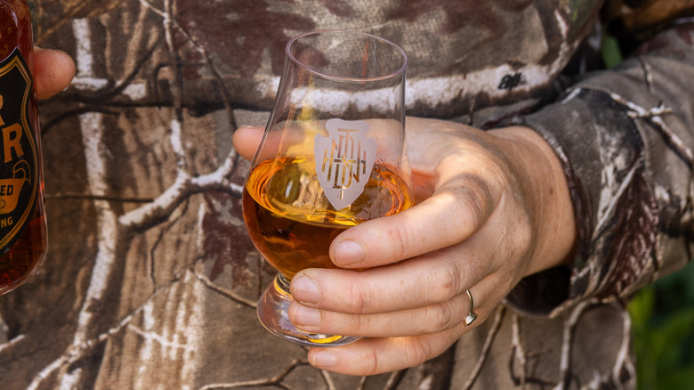 Glass of whiskey in the hand of a person in a long-sleeved camouflage shirt.