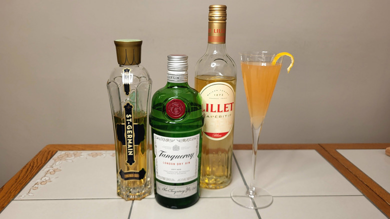 Bottles of St. Germain, Tanqueray, and Lillet Blanc next to a champagne flute containing an orange cocktail with lemon rind.