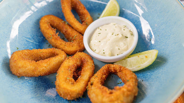 Deep-fried calamari rings beside a dish of tartar sauce on a blue plate with lime wedges