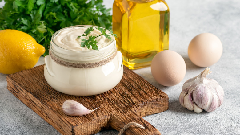 A glass jar of homemade aioli on a wooden board with ingredients all around it