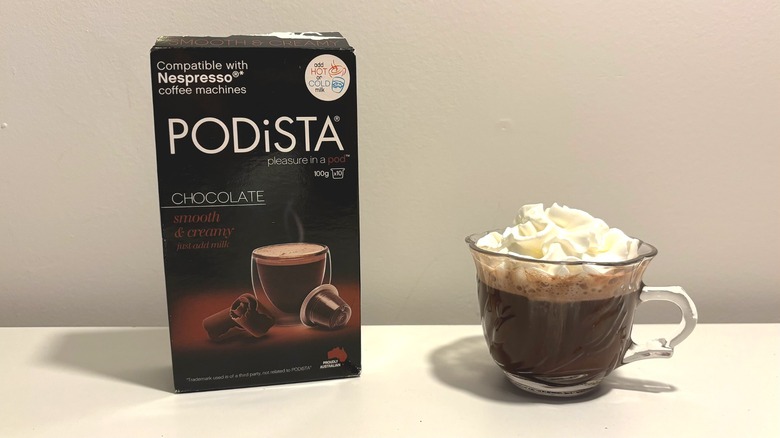Box of Podista hot chocolate pods next to clear teacup of hot chocolate on white background
