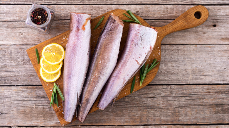 Several pieces of hake lying on a wooden chopping board next to several slices of lemon, some fresh herbs, and a jar of peppercorns