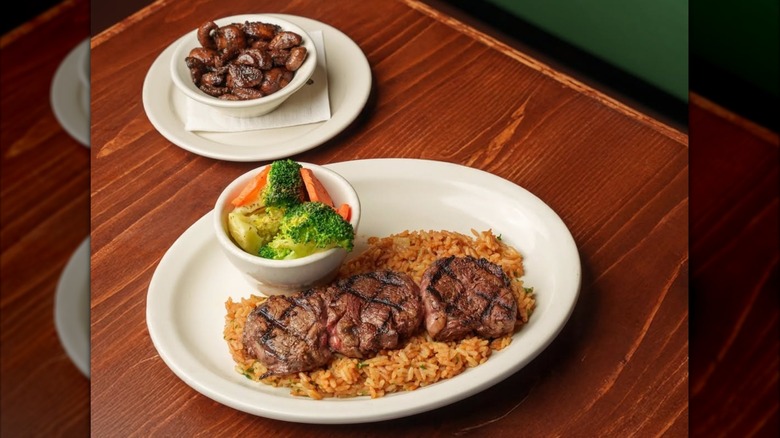 White plate of Texas Roadhouse filet medallions with steamed vegetables next to dish of sauteed mushrooms on wooden table
