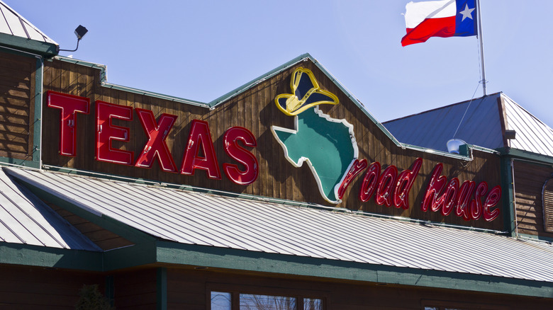 The rooftop sign of a Texas Roadhouse with a Texas flag blowing in the wind behind it