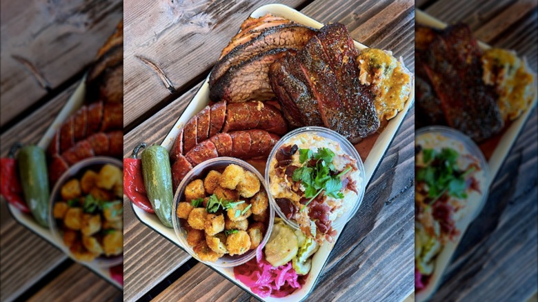 A tray of BBQ food with two steaks, sausages, fried sides, a dip, and peppers