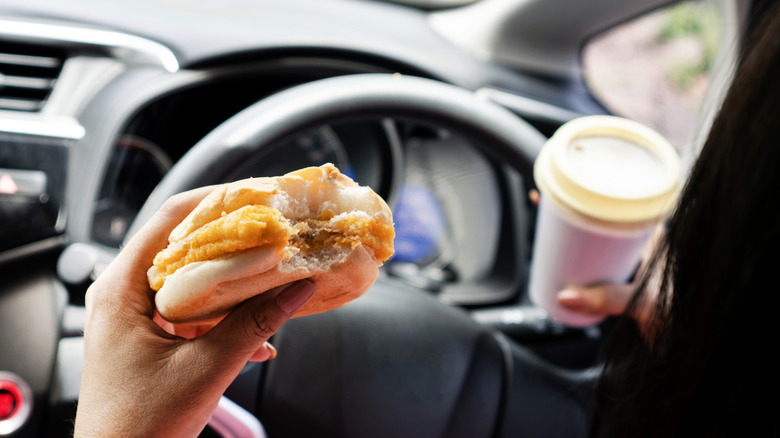 Woman driving a car while holding coffee in one hand and a breakfast sandwich in the other.