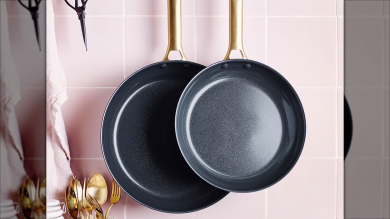 Two black and gold GreenPan Reserve frying pans hanging from a pink tile wall with gold utensils