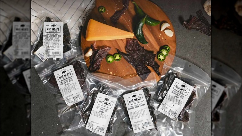 M&S Meats Montana Beef Jerky in plastic packages next to round wooden board holding wedge of cheese, jalapeno peppers, and beer jeky pieces