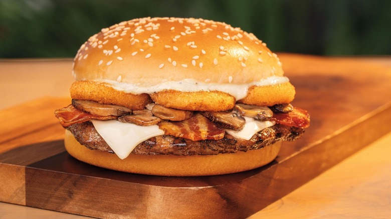 Hamburger with a sesame seed bun, cheese, bacon, mushrooms, and onion rings on a wooden cutting board