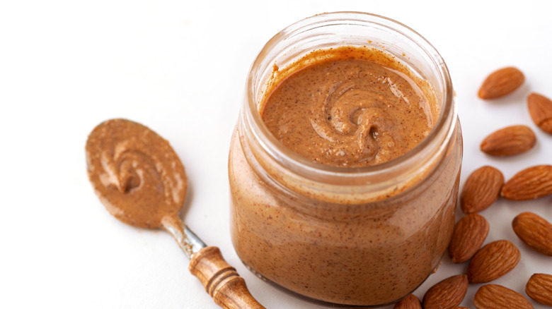 Jar of almond butter on white background, next to spoon and whole almonds