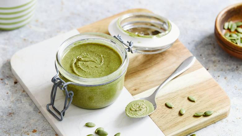 Open jar of pumpkin seed butter on wooden board with spoon and pumpkin seeds